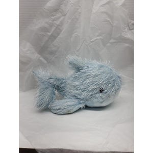 May include: A light blue plush toy whale with a soft, fuzzy texture. The whale has small black eyes and fins. The toy is lying on a white surface.