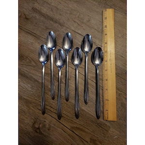 May include: Seven shiny, silver spoons with long handles are arranged on a wooden surface. A wooden ruler is placed next to the spoons. The spoons are approximately 18 cm long.