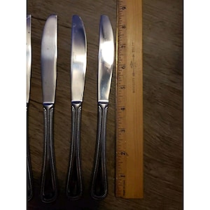 Könnte beinhalten: Ein Set aus vier Tafelmessern aus Edelstahl mit dekorativen, perlenbesetzten Griffen. Ein Holzlineal liegt neben den Messern und misst etwa 23 cm.