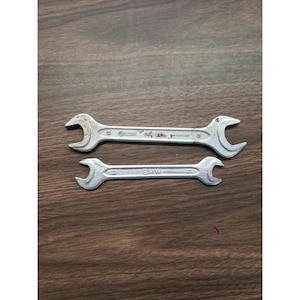 Pode incluir: Duas chaves de fenda prateadas, uma maior que a outra, são exibidas em uma superfície de madeira. A chave maior tem texto difícil de ler. A chave menor tem a palavra "BMW" impressa. Ambas as chaves têm um design curvo e aberto.