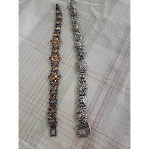 May include: Two decorative bracelets. One is dark gray with amber-colored stones in a floral pattern. The other is silver-toned with ornate square and floral designs, also with amber-colored accents. Both bracelets are displayed on a patterned fabric.