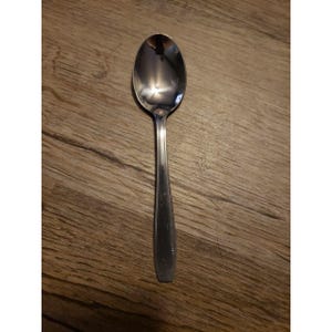 May include: A stainless steel spoon with a long, slender handle and an oval bowl. The spoon reflects the surrounding environment. The spoon is placed on a wooden surface.