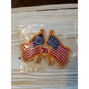 May include: Embroidered patch featuring two American flags with red and white stripes and blue star fields. A shield in the center displays the number "76" in red. The patch is in a clear plastic package.