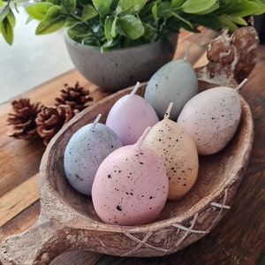 May include: A wooden bowl filled with six speckled pastel egg-shaped candles. The candles are arranged in a circle and have a white wick.