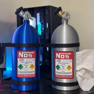 Puede incluir: Dos tanques de sistema de óxido nitroso, uno azul y otro plateado, con soportes negros. Cada tanque tiene un medidor y el logotipo "NOS" con etiquetas de advertencia. Los tanques están sobre una superficie negra.