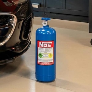 Könnte beinhalten: Eine blaue NOS-Lachgasflasche mit roten und weißen Etiketten steht neben einem schwarzen Sportwagen. Die Flasche hat ein silbernes Ventil und befindet sich in einer Garage mit einer schwarzen Werkzeugkiste.