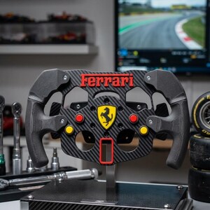 May include: A replica carbon fibre Ferrari steering wheel, with the Ferrari logo in red, yellow, and black. The wheel has red and yellow buttons and is mounted on a stand. The background includes tools and a monitor displaying a race track.