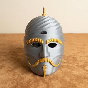 May include: A gray mask with gold accents, featuring a stylized face with eye holes, a mustache, and a pointed chin. The top of the mask has a spiraled design and a gold finial. The mask is likely for costume or decorative purposes.