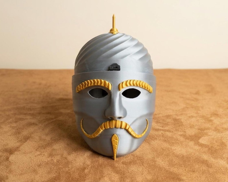 May include: A gray mask with gold accents, featuring a stylized face with eye holes, a mustache, and a pointed chin. The top of the mask has a spiraled design and a gold finial. The mask is likely for costume or decorative purposes.