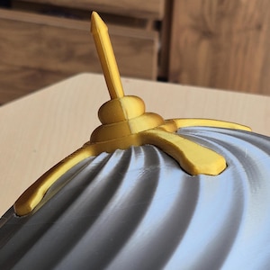 May include: Close-up of a decorative object with a gray, ridged surface and gold-colored accents. The object has a central, vertical spike and curved, wing-like extensions. The design is intricate and appears to be 3D-printed.