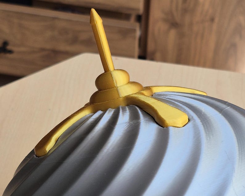 May include: Close-up of a decorative object with a gray, ridged surface and gold-colored accents. The object has a central, vertical spike and curved, wing-like extensions. The design is intricate and appears to be 3D-printed.
