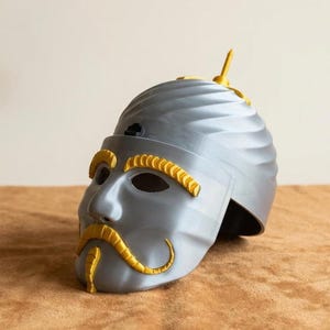 May include: A gray helmet with a face design, featuring yellow accents for the eyebrows, mustache, and a top fin. The helmet has a textured surface and is likely a costume or decorative item.