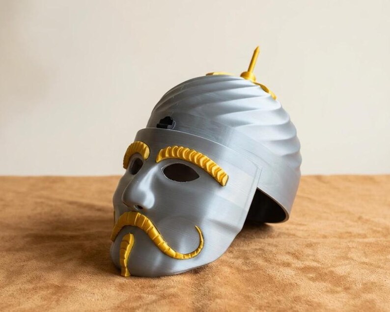 May include: A gray helmet with a face design, featuring yellow accents for the eyebrows, mustache, and a top fin. The helmet has a textured surface and is likely a costume or decorative item.