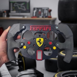 May include: A black and grey Ferrari steering wheel with red and yellow detailing. The wheel displays the Ferrari logo and text. The background includes tyres and a computer screen.