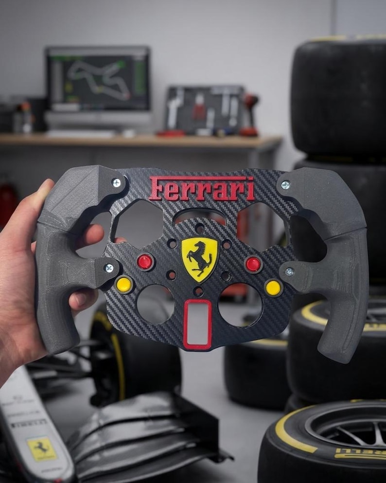 May include: A black and grey Ferrari steering wheel with red and yellow detailing. The wheel displays the Ferrari logo and text. The background includes tyres and a computer screen.