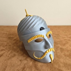 May include: A silver-colored mask with yellow accents, featuring a stylized face with eye and mouth openings. The mask has a turban-like top with a small, decorative element and a mustache.