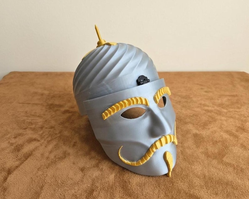 May include: A silver-colored mask with yellow accents, featuring a stylized face with eye and mouth openings. The mask has a turban-like top with a small, decorative element and a mustache.