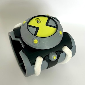 May include: A gray and black toy wristwatch with a yellow face and accents. The watch has a circular face with a yellow center and a black X design. The band is gray and black with white details.