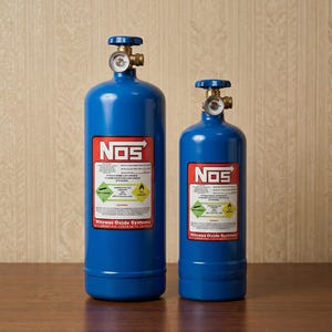May include: Two blue NOS nitrous oxide tanks of different sizes. Each tank has a red and white label with the NOS logo and safety information. The tanks have a silver valve and gauge at the top. The tanks are on a wooden surface.