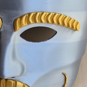 May include: A close-up of a silver and gold mask. The mask has a smooth, metallic finish with gold accents around the eyes and mouth. The eye holes are dark, and the overall design is symmetrical.