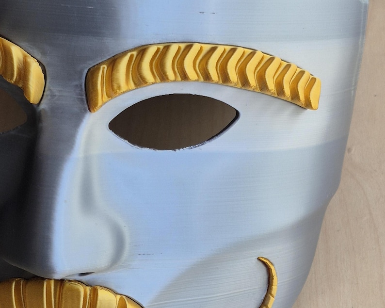 May include: A close-up of a silver and gold mask. The mask has a smooth, metallic finish with gold accents around the eyes and mouth. The eye holes are dark, and the overall design is symmetrical.