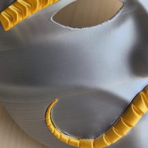 May include: A silver-colored mask with a smooth, metallic finish. The mask features two eye holes and is accented with bright yellow, curved embellishments along the edges. The design suggests a theatrical or decorative purpose.