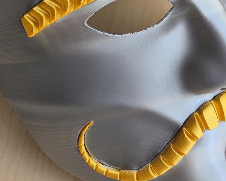 May include: A silver-colored mask with a smooth, metallic finish. The mask features two eye holes and is accented with bright yellow, curved embellishments along the edges. The design suggests a theatrical or decorative purpose.