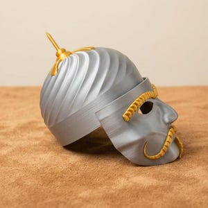 May include: A silver-colored helmet with gold accents, featuring a stylized face mask. The helmet has a textured, ridged design and a gold spike on top. The mask includes eye holes and a gold mustache.