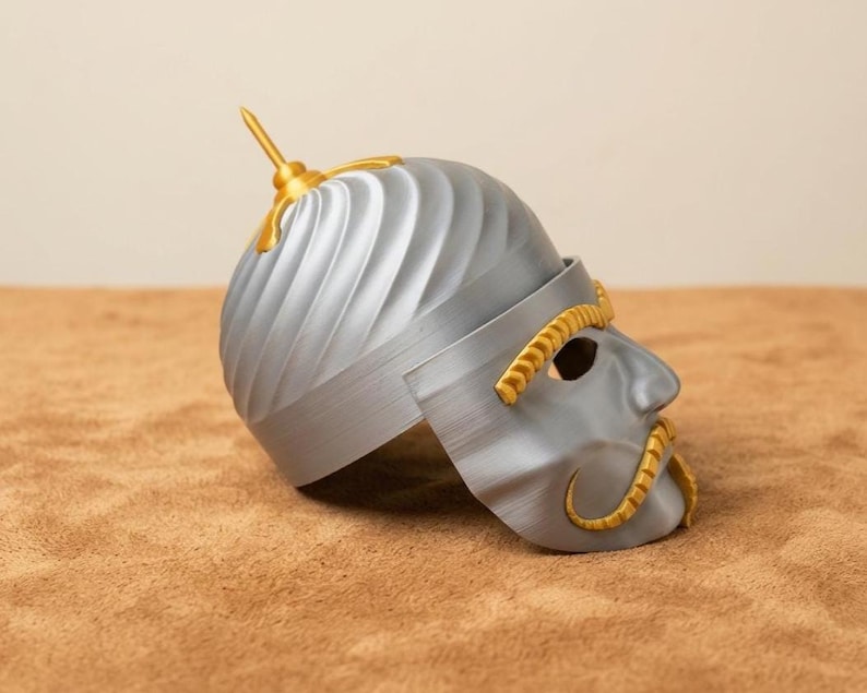 May include: A silver-colored helmet with gold accents, featuring a stylized face mask. The helmet has a textured, ridged design and a gold spike on top. The mask includes eye holes and a gold mustache.