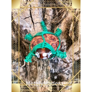 Puede incluir: Una pequeña tortuga verde y marrón hecha de hilo y una cuenta de vidrio. La tortuga está posada en una rama.