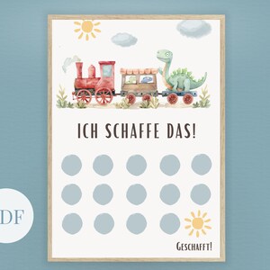 May include: A framed print featuring a watercolor illustration of a red train with a dinosaur and the text "ICH SCHAFFE DAS!". The print has a light background with blue circles and a sun graphic. The word "GESCHAFFT!" is at the bottom.