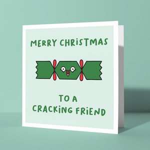 May include: A square Christmas card with a green cartoon cracker illustration and the text "MERRY CHRISTMAS TO A CRACKING FRIEND" in green. The card has a white border and is set against a light green background.