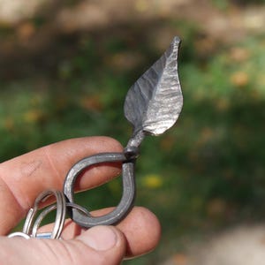 Può includere: Un portachiavi in metallo forgiato a mano con un ciondolo a forma di foglia testurizzata attaccato a un anello metallico circolare. Il pezzo in ferro grigio scuro presenta segni di martellatura visibili e una finitura rustica.