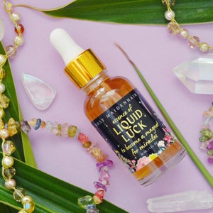 May include: A glass bottle with a gold cap and a black label that reads "essence of LIQUID LUCK to become a magnet for miracles 30 ML/1 FL OZ". The bottle is surrounded by green leaves, crystals, and colorful beads.