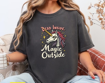 May include: Dark gray t-shirt featuring a unicorn skull graphic with the text "Dead Inside, Magic Outside". The unicorn has a gold horn, pink and purple hair, and is surrounded by stars. The shirt is a comfortable, casual style.