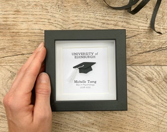 May include: A black framed picture with a white background. The picture has a black graduation cap with a white tassel and the text "University of Edinburgh" and "Michelle Tsang BSc in Psychology 2018-2022".