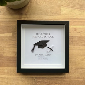 May include: A black framed picture with a white background. The picture features a black graduation cap and diploma with the text "Hull York Medical School", "Dr. Alexia Defer", "MBBS (Hons)", and "Class of 2024".