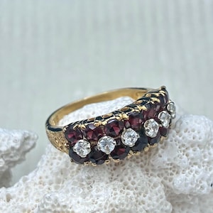 Op de afbeelding: Een gouden ring met een ontwerp van meerdere rijen met afwisselend donkerrode granaatstenen en heldere witte stenen. De band heeft een textuurafwerking aan de zijkanten en de stenen zijn in een gebogen, gelaagde opstelling geplaatst.