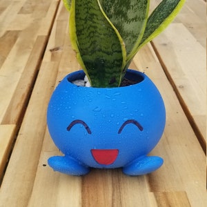 May include: A blue ceramic planter shaped like a cartoon character with a smiling face and red mouth. The planter has a small snake plant growing inside.
