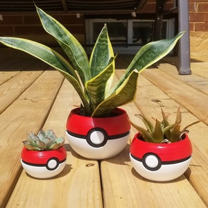 Può includere: Tre piante in vaso in vasi Pokeball rossi e bianchi. Il vaso più grande ha una pianta serpente, il vaso centrale ha una succulenta e il vaso più piccolo ha una piccola succulenta.