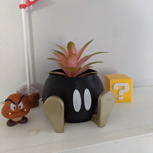 May include: A black and gold planter shaped like a character from the Super Mario Bros. video game series. The planter has two white eyes and is holding a pink succulent plant. A yellow question mark block is in the background.
