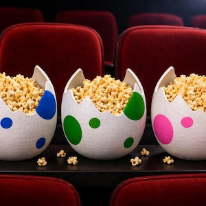 May include: Three white popcorn bowls shaped like cracked eggs, each with a different color of polka dots: blue, green, and pink. The bowls are filled with popcorn and sit on a tray in a movie theater.
