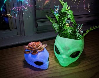 Puede incluir: Dos macetas con forma de alienígena y superficies lisas. La maceta blanca más pequeña contiene una suculenta de color cobre metálico. La maceta verde brillante más grande contiene un helecho y pequeñas figuras alienígenas iluminadas.