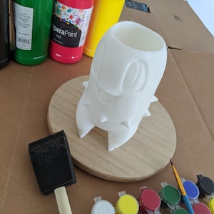 Pode incluir: Um vaso de cerâmica branco em forma de fantasma com um rosto feliz, sentado em uma base de madeira. O vaso é cercado por materiais de pintura, incluindo um pincel de esponja, um pincel e pequenos recipientes de tinta de várias cores, incluindo vermelho, amarelo, verde, azul, preto e branco.