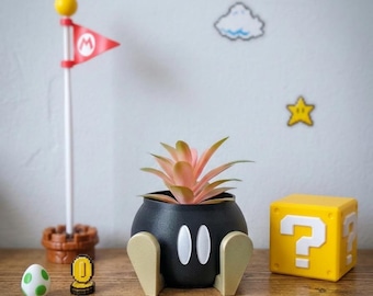 May include: A black, spherical planter shaped like a Bob-omb character with white oval eyes and gold feet, containing a small pink and green succulent. Beside it are a Mario flag, a small white egg with green spots, a gold coin, and a yellow question block.