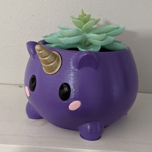 Può includere: Vaso per piante a forma di unicorno viola con un corno dorato e occhi neri. Il vaso contiene una piccola pianta grassa.