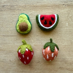 Puede incluir: Cuatro adornos de fruta de fieltro: un aguacate verde, una rodaja de sandía roja, una fresa roja y una fresa naranja.