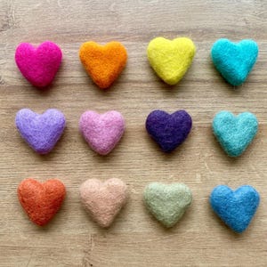 May include: A collection of twelve felt heart-shaped ornaments in various colors, including pink, orange, yellow, teal, purple, and blue. The hearts are arranged on a light brown wooden surface, showcasing their vibrant hues and textured appearance.