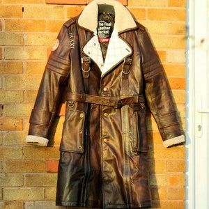 May include: A brown leather jacket with a fur collar and a belt. The jacket has a zipper closure and multiple pockets. The jacket has a patch on the left sleeve that says "The Real Leather Jacket".