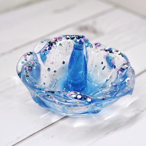May include: A blue ring holder with a wavy edge and a clear center filled with colorful glitter. The holder has a blue ring stand in the center.